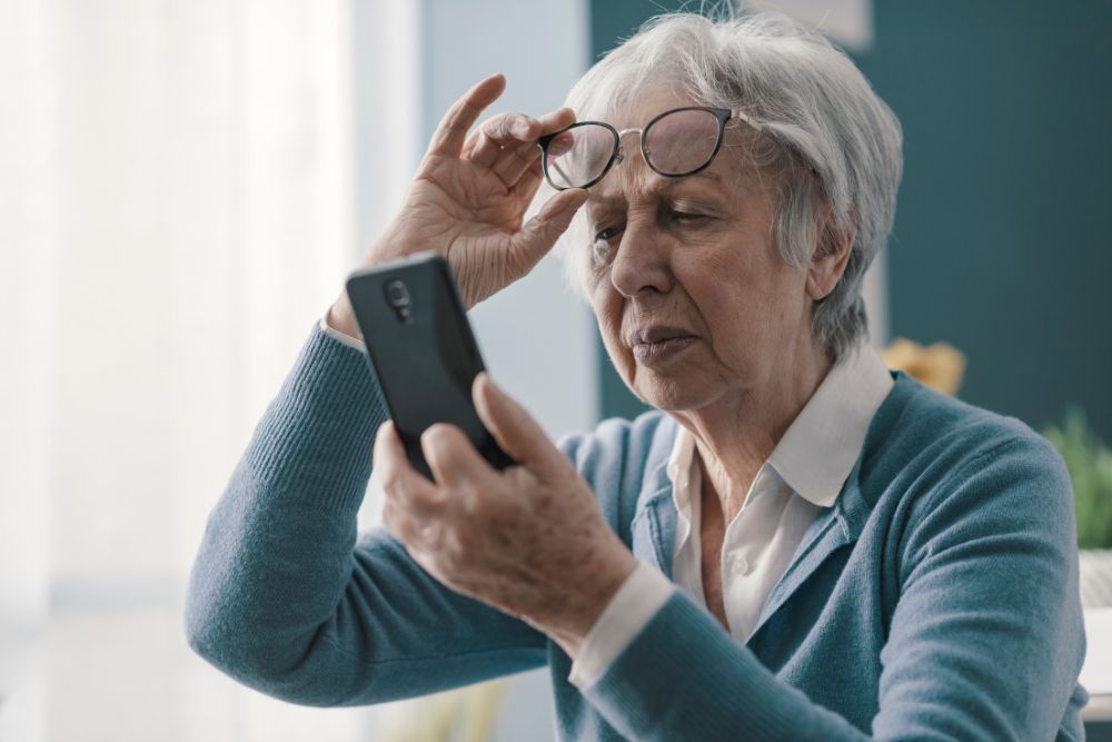 Older woman holding glasses while looking at a smartphone, representing blurred vision symptoms